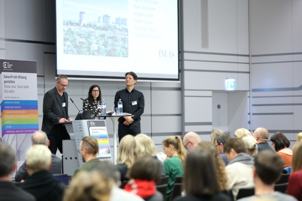 Drei Personen am Rednerpult vor Publikum