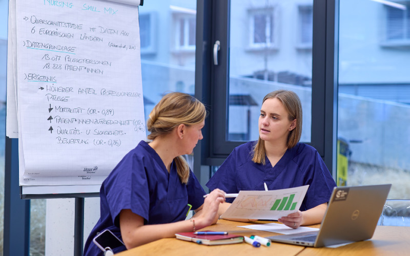 2 PFlegepersonen vor einem Laptop, hinter ihnen ein Flipchart mit Bulletpoints