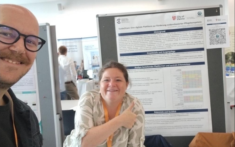Selfie von zwei Personen vor einem an einer Pinwand angebrachten Poster mit der Beschreibung von Guide2Care