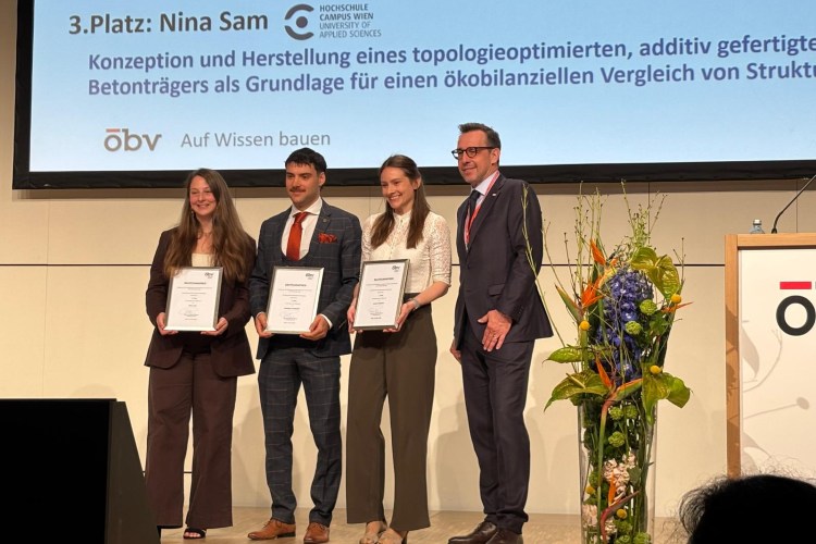 Vier Personen stehen auf einer Bühne und halten Urkunden; im Hintergrund ist eine Präsentationsfolie mit dem 3. Platz und einem Projekttitel zu sehen, daneben ein Blumenarrangement.