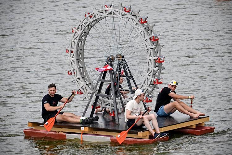 © InformationsZentrum Beton Vier Personen sitzen auf einem schwimmenden Floß und paddeln auf einem Gewässer. In der Mitte des Floßes befindet sich ein großes, detailreich nachgebildetes Wiener Riesenrad aus Beton. Die Konstruktion dient als ungewöhnliches Wasserfahrzeug und ist Teil eines Wettbewerbs. Im Hintergrund ist die Wasseroberfläche zu sehen.