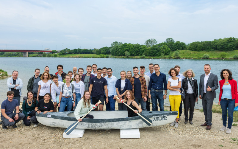 Frauen und Männer stehen hinter einem Betonkanu, zwei Frauen sitzen mit Paddeln darin