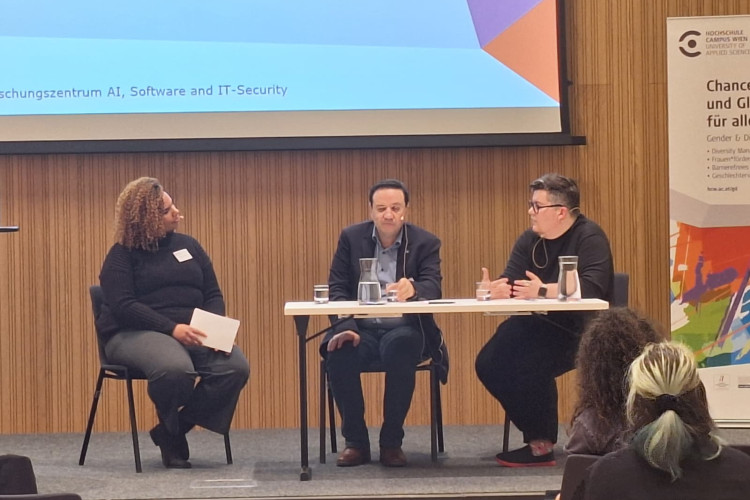 © Kristina Zimmermann Drei Personen sitzen auf einer Bühne an einem Tisch und diskutieren vor Publikum. Im Hintergrund ist eine Präsentationsfolie des Forschungszentrums AI, Software and IT-Security zu sehen.