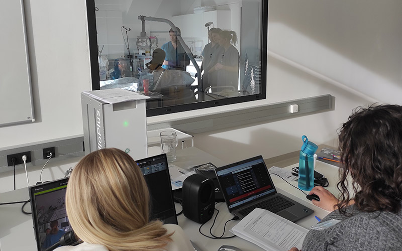 © HCW / Ströhle Zwei Frauen vor Bildschirmen und einer Glasscheib, im dahinter befindlichen Raum wir ein Schauspielpatient von Studierenden monitored