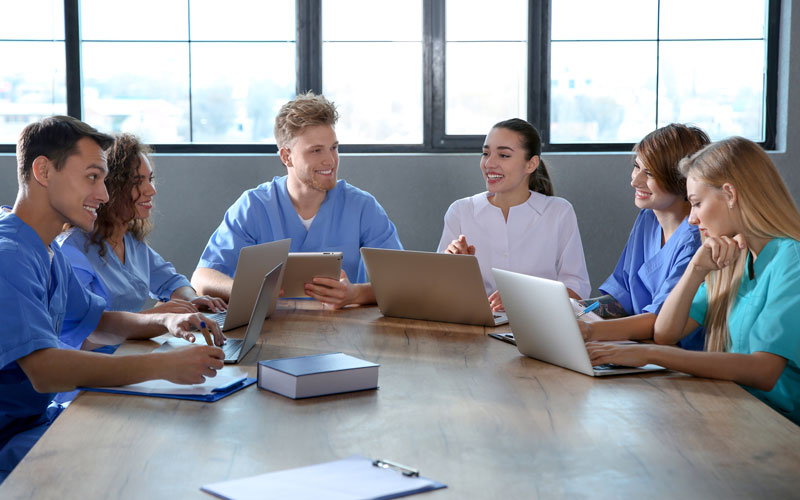 Personen in typischer Pflegeberufskleidung sitzen um einen Tisch, eine Person trägt etwas vor.