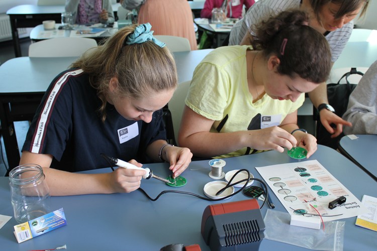 Drei Jugendliche sitzen an einem Tisch und arbeiten konzentriert mit Lötkolben an kleinen elektronischen Bauteilen. Vor ihnen liegen Werkzeuge, Kabel und Platinen.