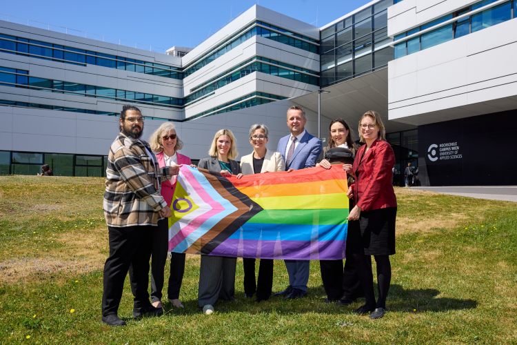 © HCW/Schedl Sechs Personen stehen vor dem Gebäude der Hochschule Campus Wien auf einer Wiese und halten gemeinsam eine Progress-Pride-Flagge. Im Hintergrund ist die moderne Hochschulfassade bei sonnigem Wetter zu sehen. Die Gruppe blickt in die Kamera und setzt ein sichtbares Zeichen für Vielfalt und Inklusion.