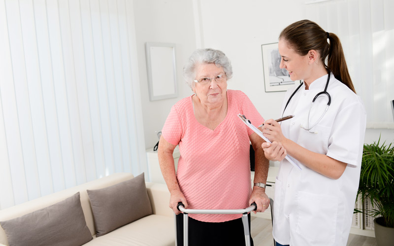 © iStock/ JPWALLET Junge Dame in weißem Kittel mit Stift und Klemmbrett neben älterer Dame an Gehhilfe